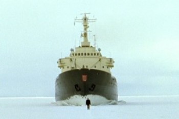 A screenshot of a video artwork. It depicts a man walking on the ice with a large ship not far behind him, breaking through the ice on which the man just walked.