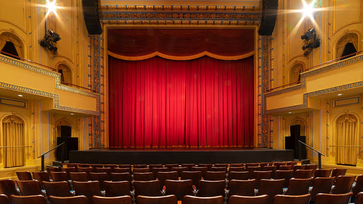A photo of a theater where actors perform.  There are gold walls and a red curtain.
